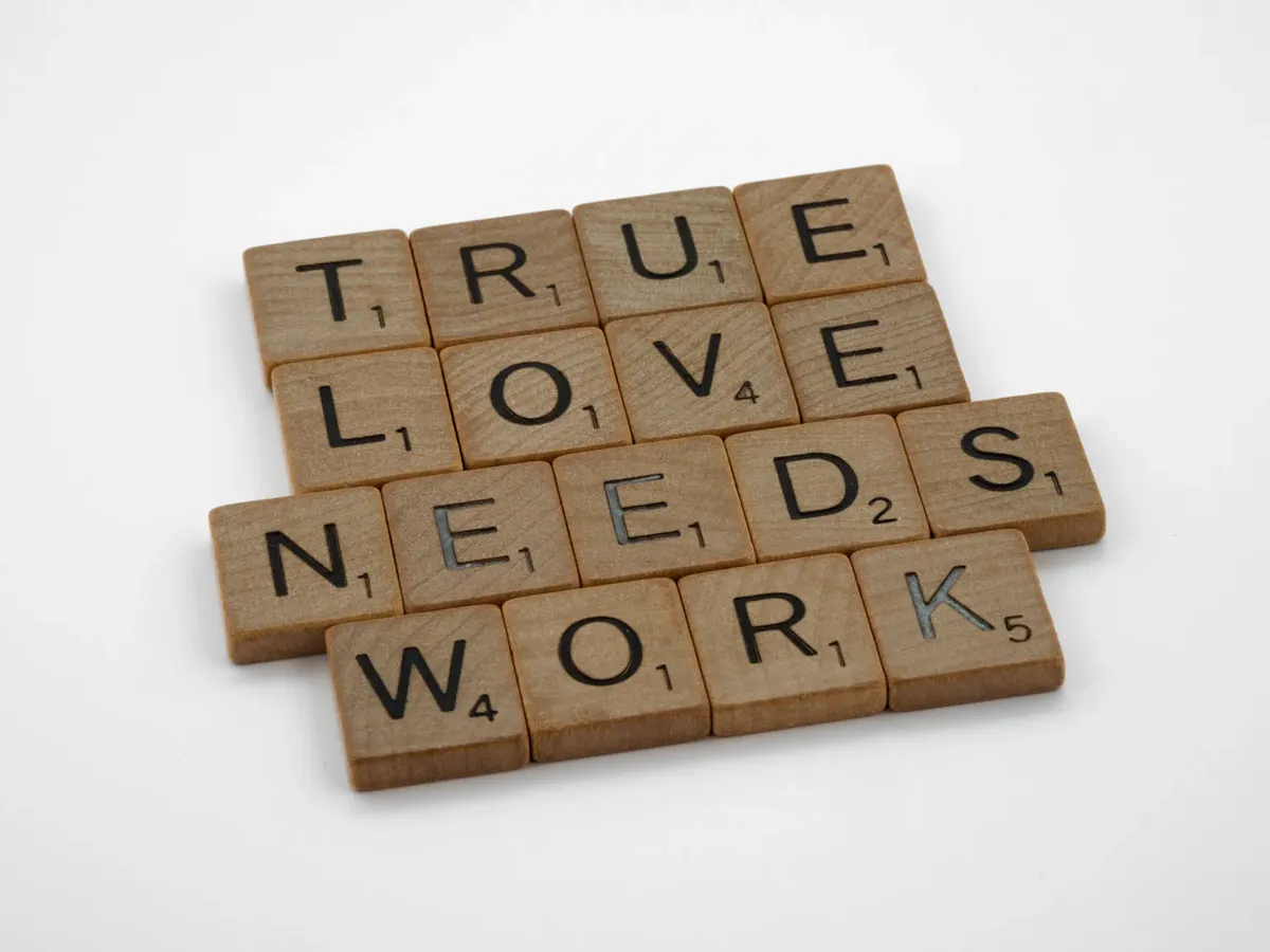 A close-up of wooden Scrabble tiles on a white surface spelling 'True Love Needs Work'.