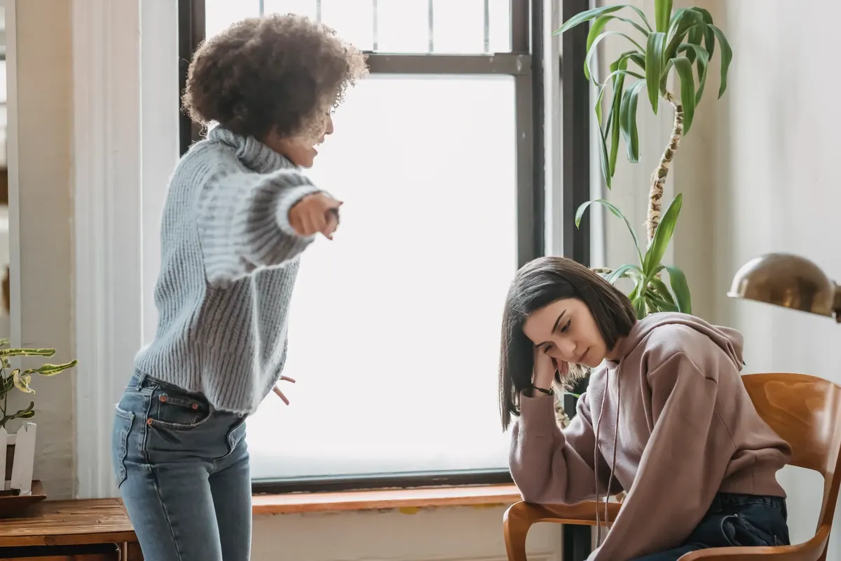 Angry African American woman pointing away while looking at unhappy female while having conflict in light room with green plant at home
